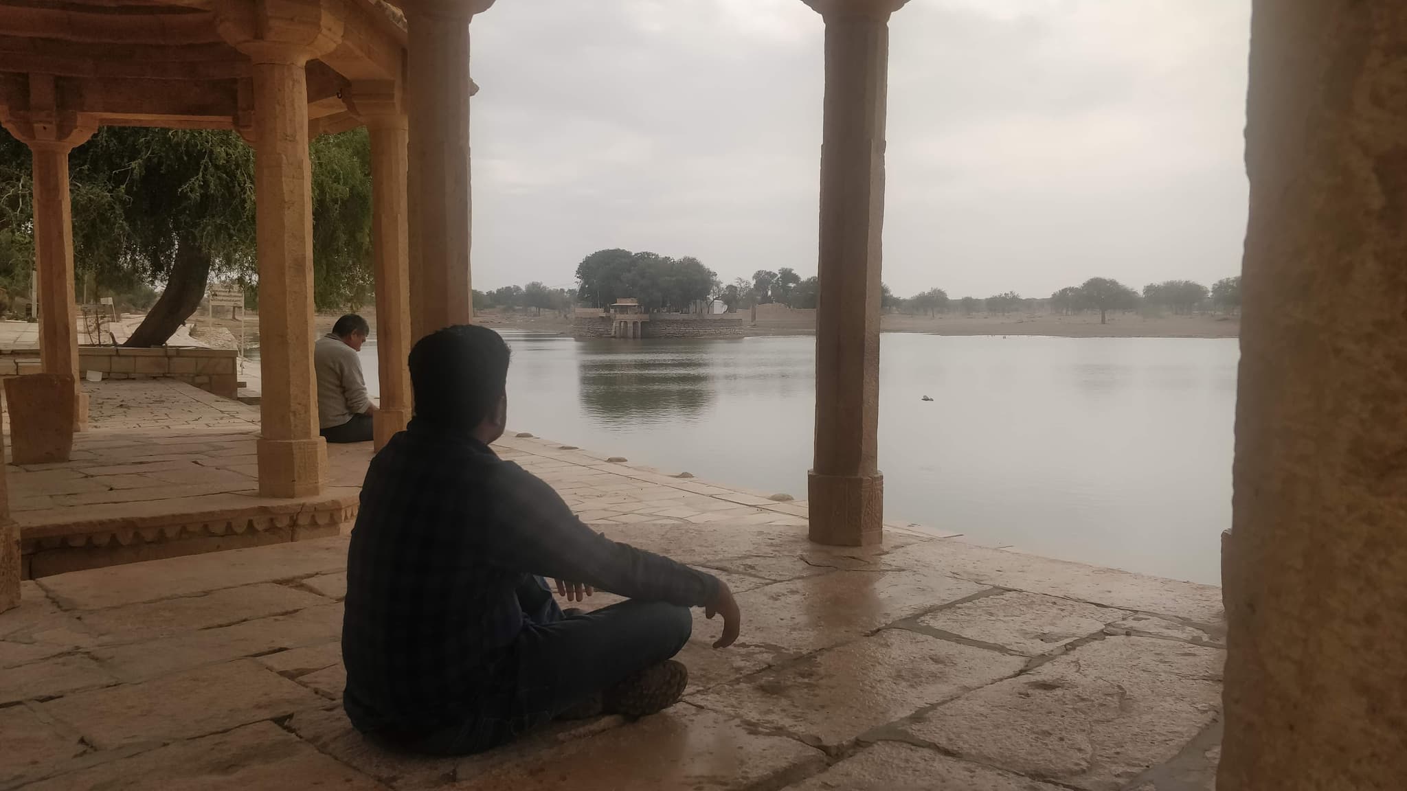 Person sitting cross-legged in ornate sandstone pavilion overlooking Gadisar Lake