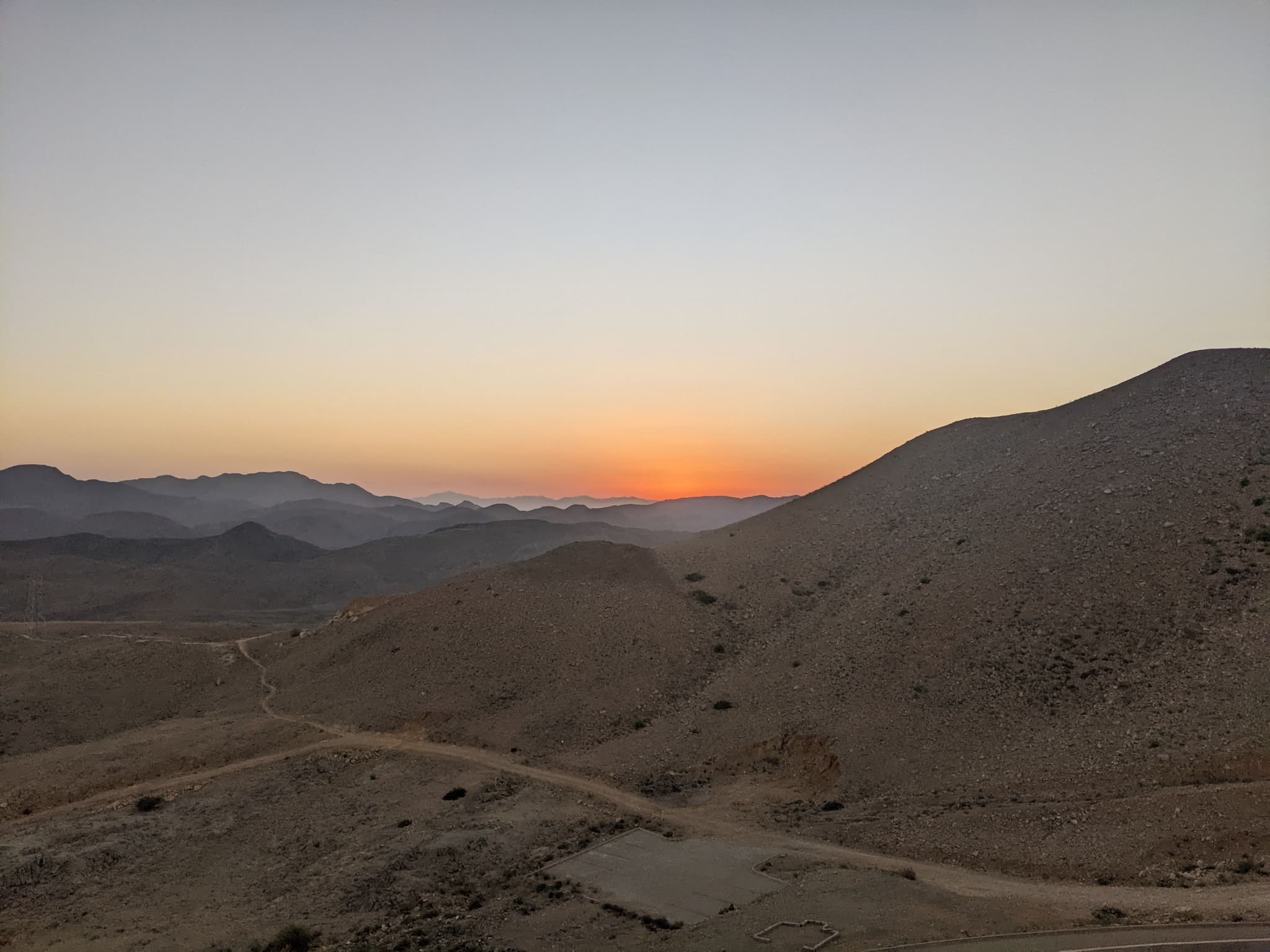 Sunset over barren desert mountains, orange-gold gradient sky, winding road in valley