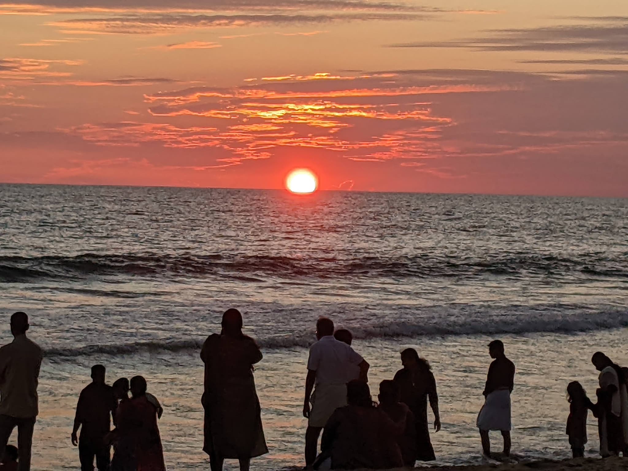 Vivid red-orange sunset with sun as bright orb over dark sea, silhouetted figures