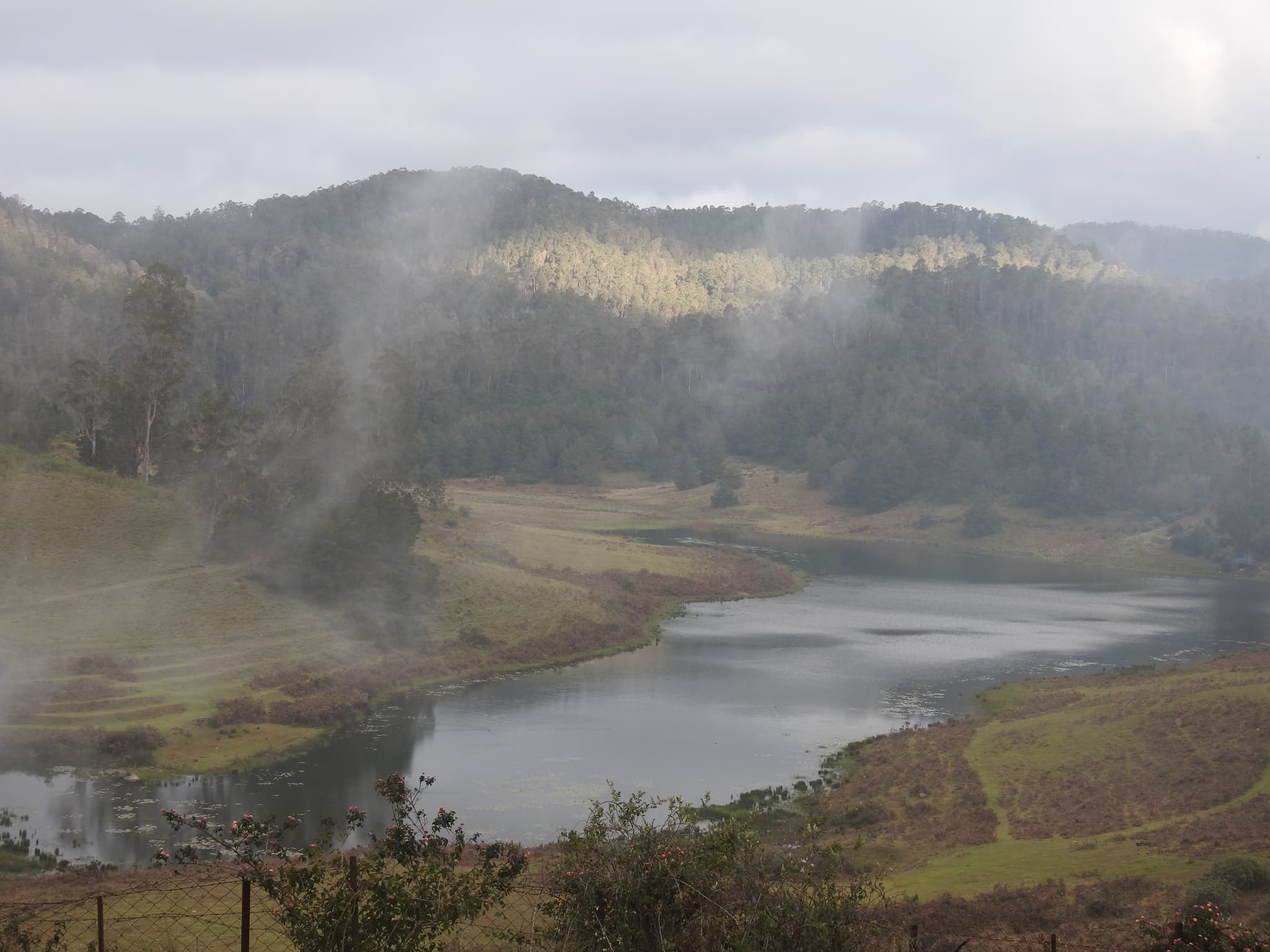 Misty lake with forested hills, low clouds, and light fog rising from water