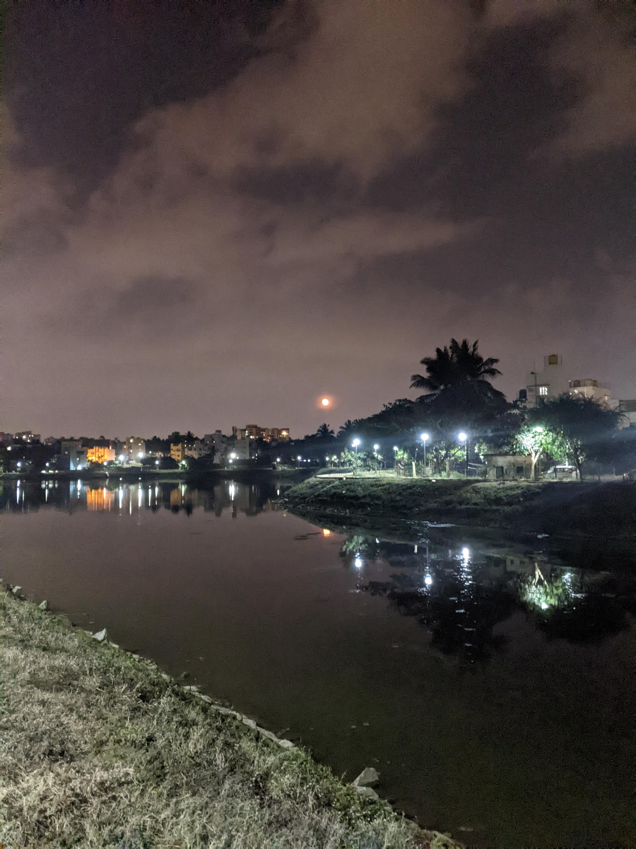 Night reflection of city lights on calm lake with orange moon rising through clouds