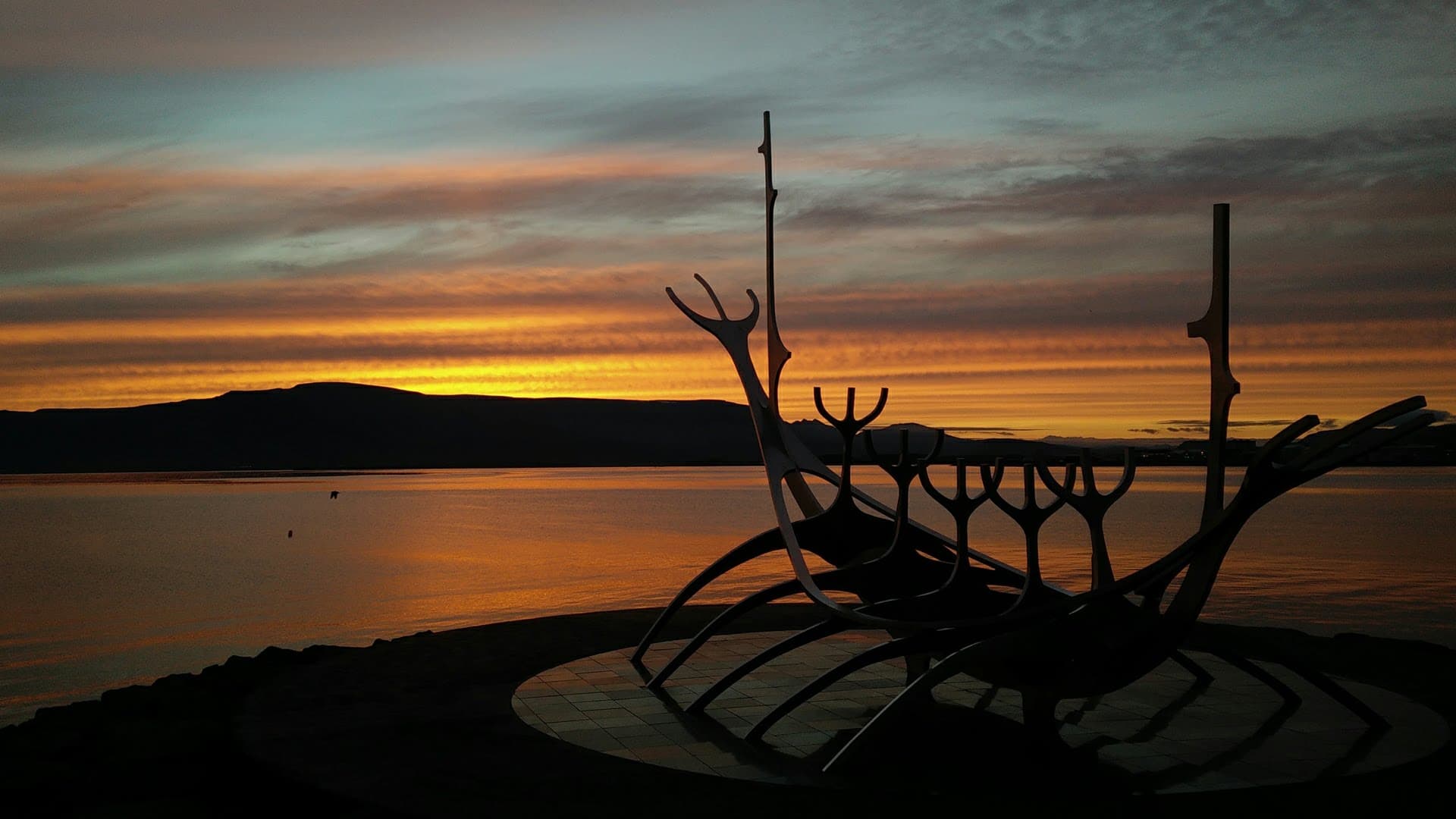Sun Voyager sculpture silhouetted against dramatic orange-gold sunset over water