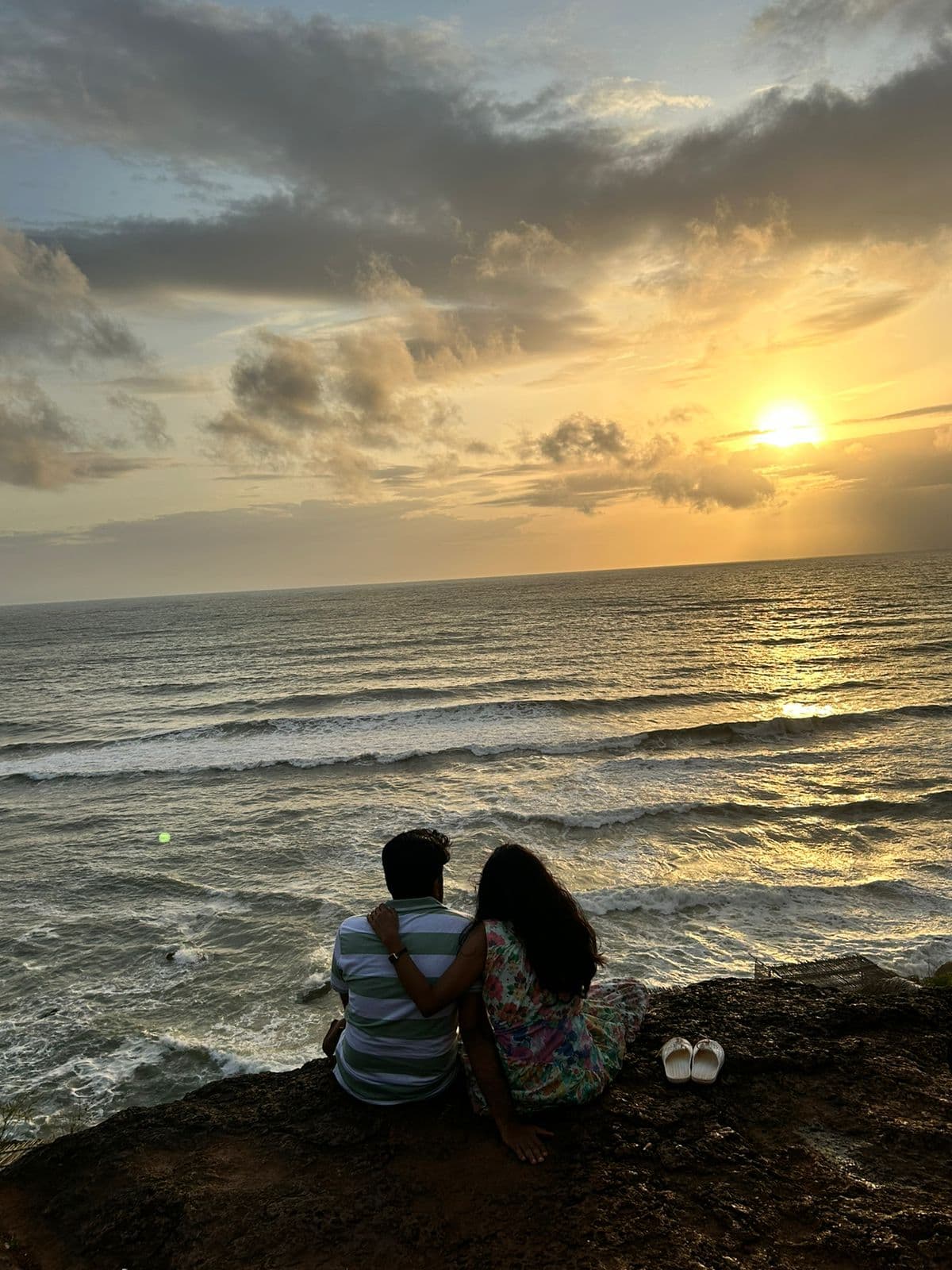 A couple sits on a cliff, silhouetted against a vibrant sunset over the ocean, with the sun reflecting on the water's surface.